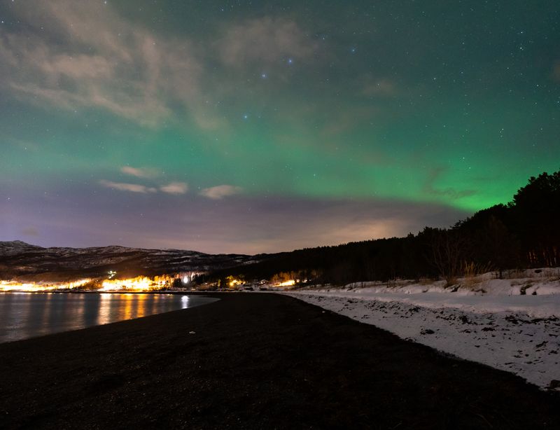 Green northern lights over a snowy fjord shore, with town lights along the water