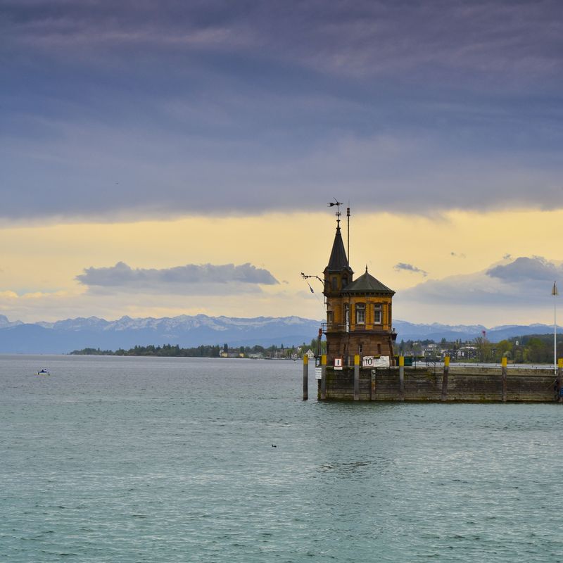 Lakeside lighthouse tower on a pier, snowy Alps on the horizon